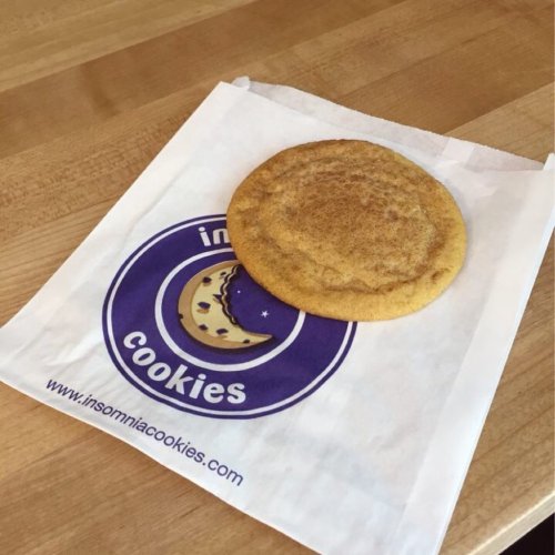 A single cookie on a branded paper bag from Insomnia Cookies, resting on a wooden surface.
