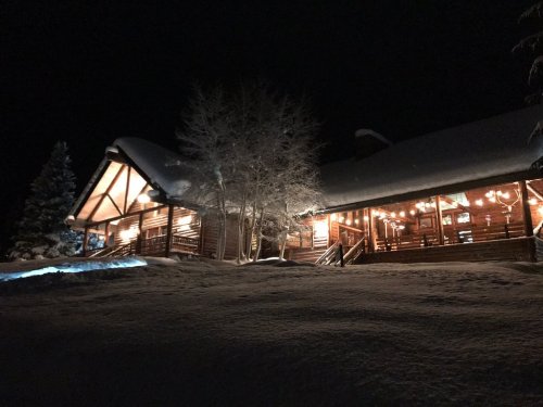 A cozy log cabin illuminated at night, surrounded by snow and trees, creating a warm winter atmosphere.