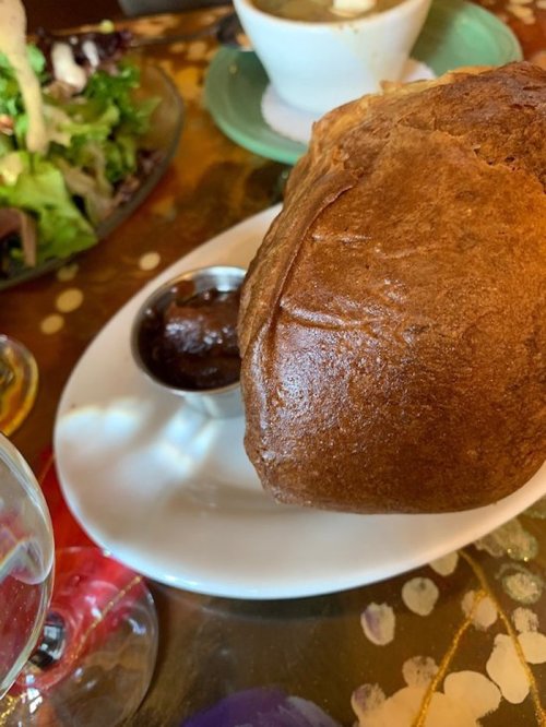 A freshly baked bread roll on a plate with a small dish of sauce, alongside a salad and soup in the background.