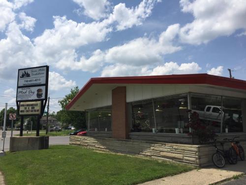 A diner with a red roof and large windows, surrounded by green grass and a cloudy sky. A sign displays contact info.