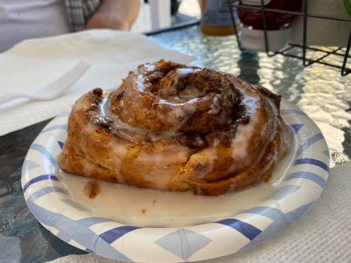 A cinnamon roll on a paper plate, drizzled with icing, resting on a glass table.