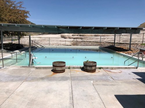 A partially filled outdoor pool with a covered area, surrounded by concrete and two barrels, under a clear blue sky.