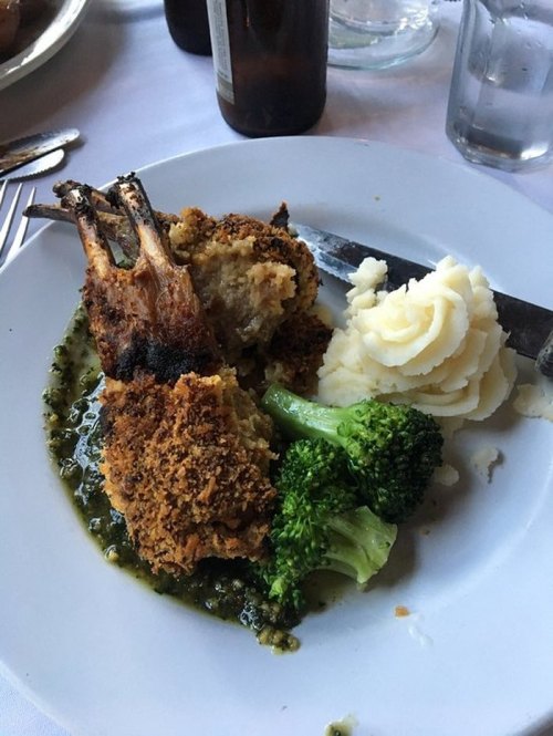 A plate with two breaded lamb chops, mashed potatoes, broccoli, and a green sauce on a white tablecloth.