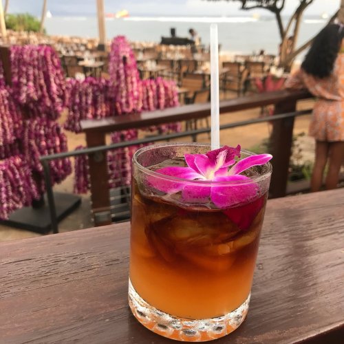 A tropical cocktail garnished with a pink flower, set against a backdrop of outdoor dining and floral decorations.