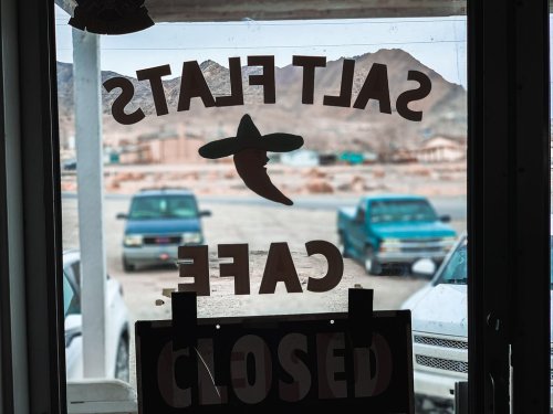 A view through a café window with "CLOSED" sign, showing parked cars and mountains in the background.