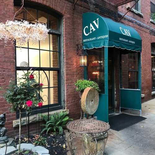 Exterior of a restaurant named "CAV," featuring a green awning, brick walls, and decorative plants.