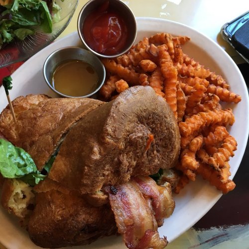 A plate featuring a sandwich with bacon and lettuce, accompanied by crinkle-cut sweet potato fries and dipping sauces.