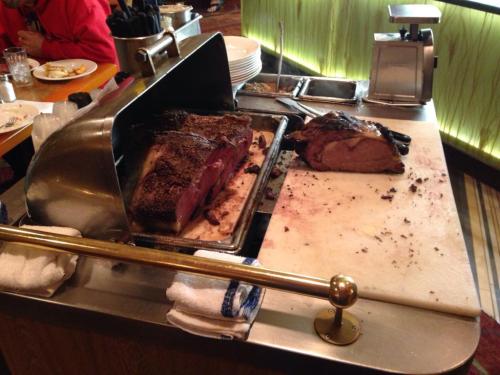 A carving station with large cuts of roasted meat on a serving cart, with utensils and plates in the background.