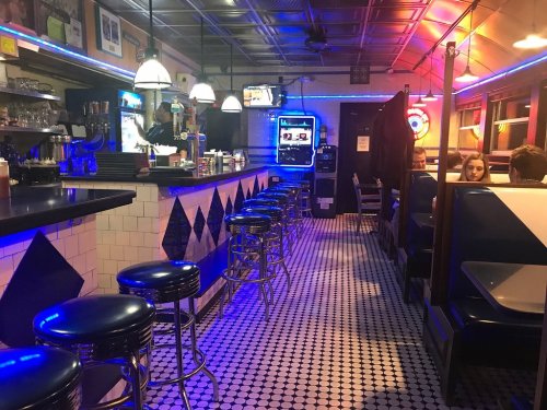 A retro diner interior with blue and pink lighting, featuring bar stools, a counter, and booths along the walls.