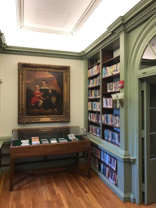 A cozy library corner featuring a large painting, wooden display case, and shelves filled with books.
