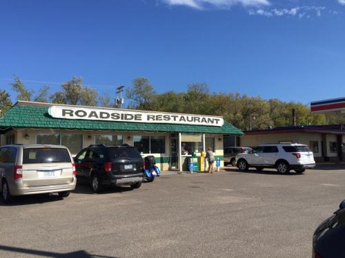 A roadside restaurant with a green roof, surrounded by parked cars and trees under a clear blue sky.