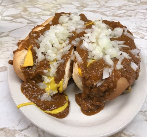 Two hot dogs topped with chili, shredded cheese, and chopped onions on a white plate.