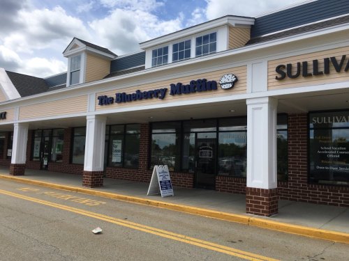 Exterior of a storefront labeled "The Blueberry Muffin" with a sign and nearby businesses in a shopping plaza.