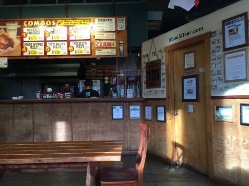 Interior of a casual restaurant with a menu board, counter, and wooden tables. Two staff members are visible behind the counter.