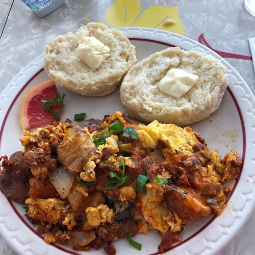 A plate with scrambled eggs mixed with vegetables and sausage, served with two buttered biscuits and a slice of grapefruit.