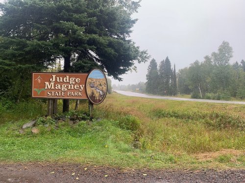 Sign for Judge Magney State Park beside a road, surrounded by trees and tall grass on a cloudy day.