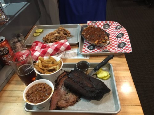 A table with trays of barbecue, including brisket, pulled pork, fries, baked beans, pickles, and drinks.