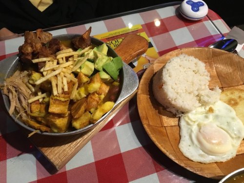 A metal bowl filled with fried plantains, avocado, and meat, alongside a wooden plate with rice and a fried egg.