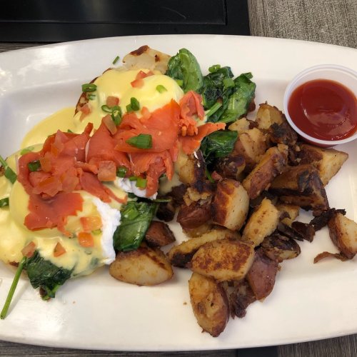 A plate of eggs Benedict with spinach, smoked salmon, diced tomatoes, and crispy potatoes, served with a side of ketchup.