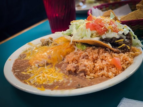 A plate of Mexican food featuring rice, refried beans, cheese, and a taco topped with lettuce and tomatoes.