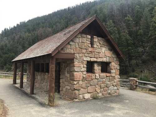 A rustic stone building with a sloped roof, surrounded by trees and mountains, along a paved path.