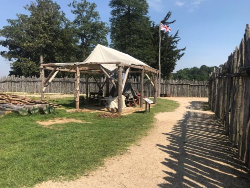 A rustic tent structure inside a wooden stockade, with a British flag flying and a clear blue sky above.