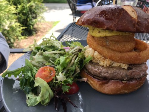 A gourmet burger with a pretzel bun, topped with onion rings and sauerkraut, served with a side salad.