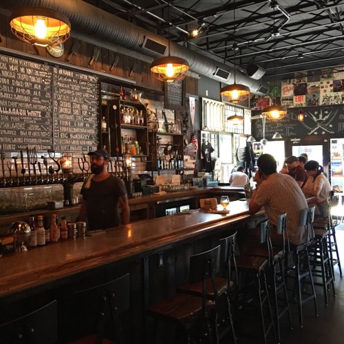 A cozy bar interior with a long wooden counter, patrons seated, and warm lighting illuminating the space.