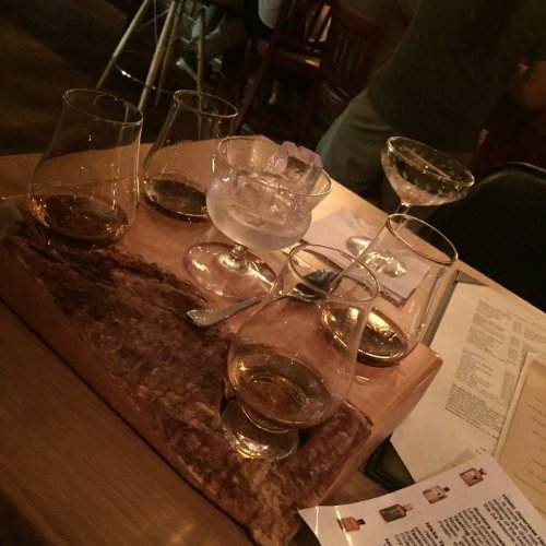 A wooden board with five glasses of whiskey and a cocktail, set on a table in a dimly lit bar.