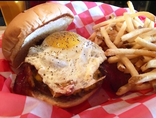 A burger with a fried egg on top, served with a side of crispy French fries in a red and white checkered basket.