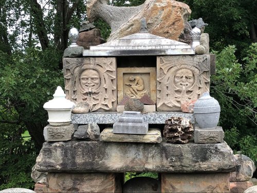 A stone structure featuring intricate carvings, sculptures, and various decorative elements, surrounded by greenery.