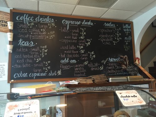 Chalkboard menu displaying prices for coffee, tea, espresso drinks, sodas, and add-ons at a café.