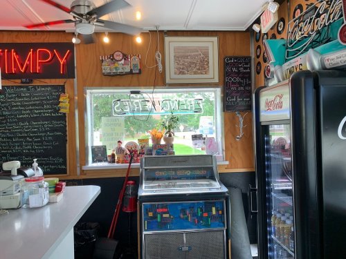 A cozy diner interior featuring a jukebox, neon signs, and a menu board, with a view of the outdoors through a window.