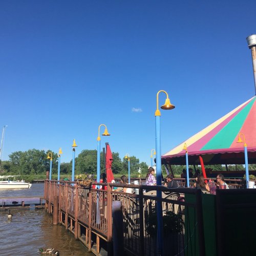 A colorful riverside restaurant with a striped canopy, people dining, and a clear blue sky above.