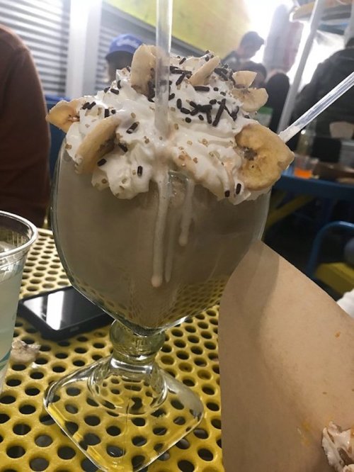 A large dessert glass filled with a creamy shake, topped with whipped cream, banana slices, and chocolate sprinkles.
