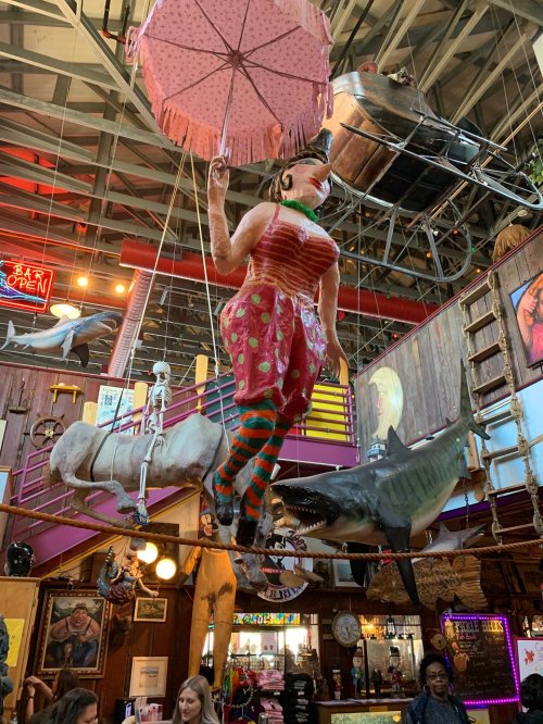A whimsical indoor scene featuring a large, colorful figure holding an umbrella, suspended above various quirky decorations.