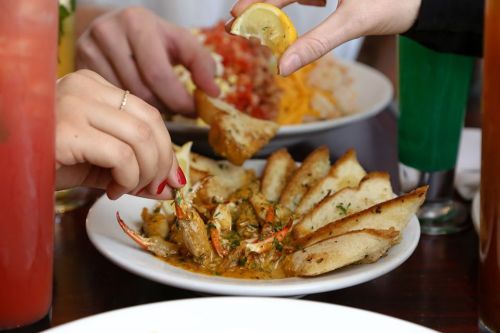 A plate of crab dish with bread, as someone squeezes lemon over it. Colorful drinks are in the background.