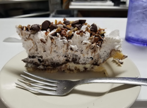 A slice of dessert pie topped with whipped cream, chocolate chips, and nuts on a white plate with a fork.