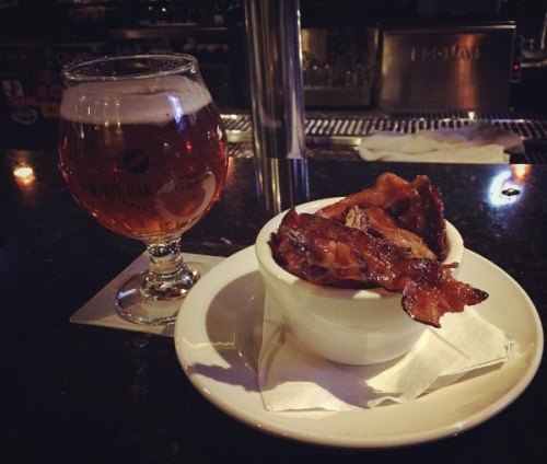 A glass of amber beer next to a small bowl of crispy bacon on a bar counter.