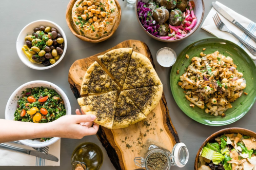 A wooden table with various Mediterranean dishes, including a sliced flatbread, salads, and bowls of olives and chickpeas.