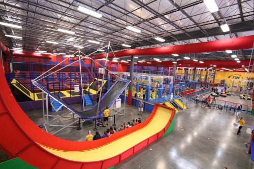 A vibrant indoor trampoline park with various play structures, slides, and climbing areas filled with people enjoying activities.
