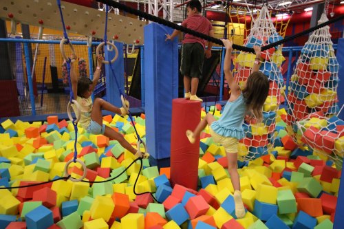 Children play in a colorful foam pit, climbing and swinging on ropes and structures in a vibrant indoor playground.