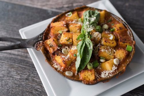 A cast-iron skillet filled with golden polenta squares, topped with basil, green onions, and grated cheese.
