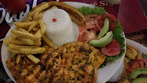 A plate of grilled chicken, rice, fries, fried plantains, and a fresh salad with tomatoes and avocado.