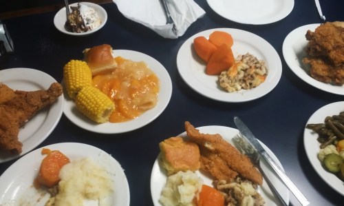A table spread with plates of fried chicken, corn, sweet potatoes, green beans, and dessert.