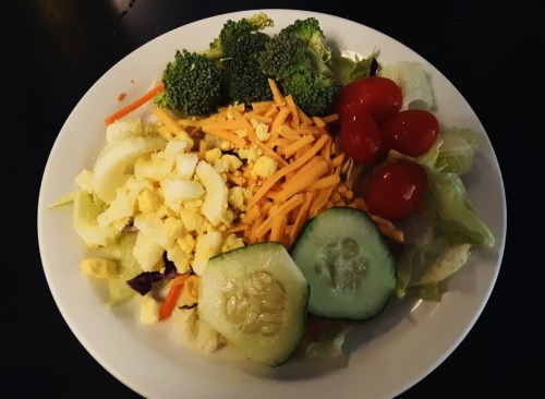 A colorful salad with lettuce, broccoli, cucumber, cherry tomatoes, shredded cheese, and chopped eggs on a white plate.