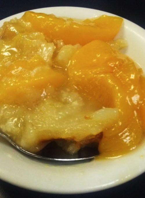 A bowl of peach cobbler with a golden peach topping and a spoon resting beside it.