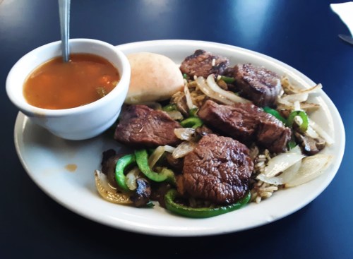 A plate of beef strips with green peppers and onions, served with rice and a side of soup and a bread roll.