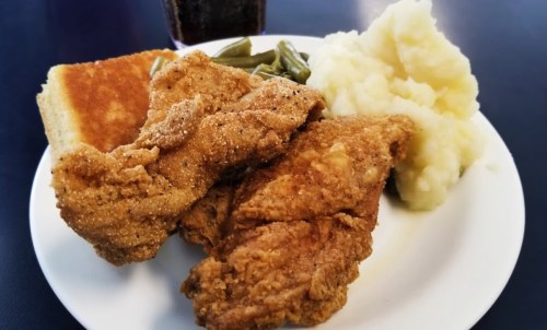 A plate of fried chicken, mashed potatoes, green beans, and cornbread, with a drink in the background.