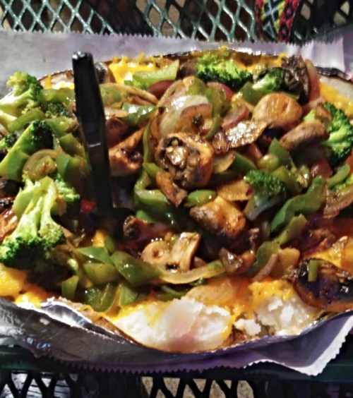 Baked potato topped with melted cheese, sautéed mushrooms, green peppers, and broccoli on a metal tray.